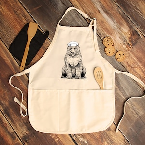 May include: Cream-colored apron with a bear wearing a chef's hat. The bear illustration is in black and white. A wooden spatula and a black oven mitt are on the left. Chocolate chip cookies are on the right. The apron has two pockets.