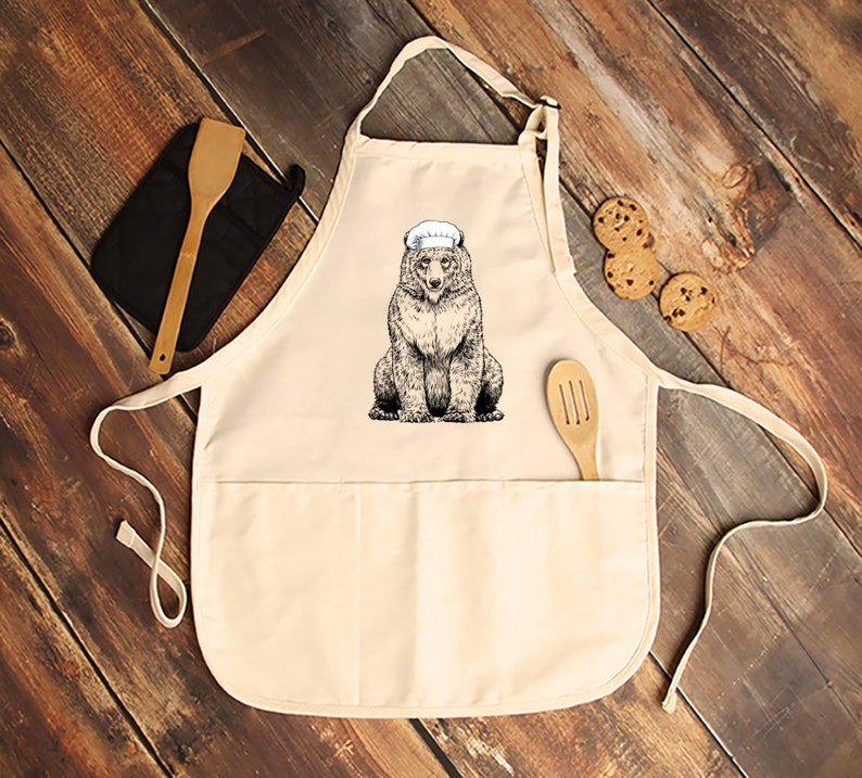 May include: Cream-colored apron with a bear wearing a chef's hat. The bear illustration is in black and white. A wooden spatula and a black oven mitt are on the left. Chocolate chip cookies are on the right. The apron has two pockets.