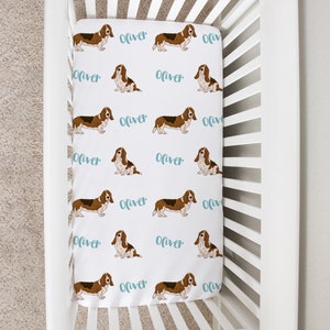 May include: White crib with a white fitted sheet featuring a repeating pattern of brown basset hounds and the name "Oliver" in light blue cursive.