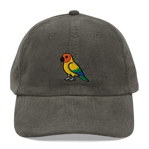 May include: A dark green corduroy baseball cap with a colorful embroidered parrot design. The parrot has a red head, yellow body, and blue and green wings. The cap has a curved brim and a fabric strap.