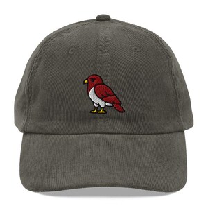 May include: A grey corduroy baseball cap with an embroidered red and white bird design. The bird has a yellow beak and is perched on the cap. The hat has a curved peak and an adjustable strap.