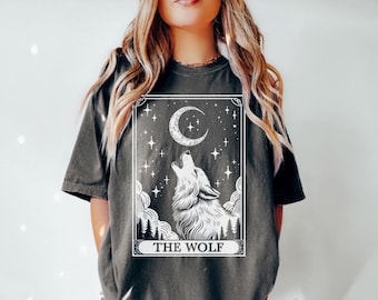May include: A dark grey t-shirt featuring a white graphic of a wolf howling at the moon. The graphic is in the style of a tarot card and includes the text "THE WOLF".