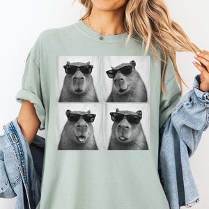 Puede incluir: Camiseta verde claro con una imagen de cuatro paneles de un carpincho con gafas de sol. El carpincho es en blanco y negro, con un fondo blanco. La camiseta es de corte holgado.