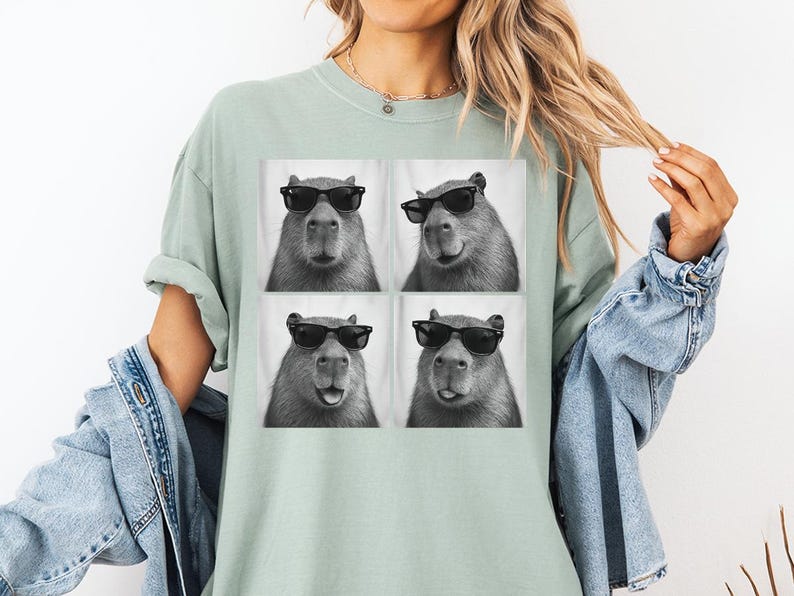Puede incluir: Camiseta verde claro con una imagen de cuatro paneles de un carpincho con gafas de sol. El carpincho es en blanco y negro, con un fondo blanco. La camiseta es de corte holgado.