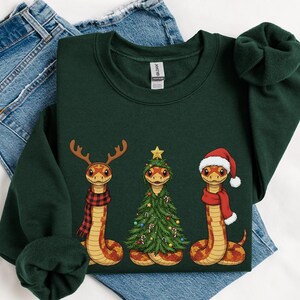 May include: Dark green sweatshirt featuring three cartoon snakes. One snake wears a reindeer antler scarf, another is a Christmas tree, and the third wears a Santa hat and scarf. The sweatshirt is displayed on top of blue jeans.