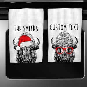 May include: Two white kitchen towels with a black and white illustration of a buffalo wearing a Santa hat and sunglasses. The text "THE SMITHS" and "CUSTOM TEXT" is printed on the towels.