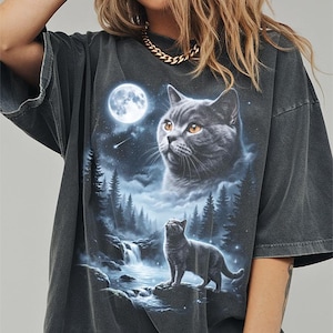 May include: Dark gray t-shirt featuring a detailed graphic of a cat under a full moon, with a waterfall and forest scene. The cat's image is in the center of the shirt, with a second cat looking up at the moon. The shirt has short sleeves.
