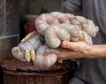 May include: A collection of hand-dyed yarn skeins in a variegated color palette of cream, beige, and soft brown with subtle speckles of deep red. The yarn is twisted into bundles and decorated with two dried wheat stalks tucked into the fibers.