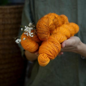 Könnte beinhalten: Eine Handvoll leuchtend orangefarbener Garnknäuel, akzentuiert mit kleinen weißen Blüten. Das Garn hat eine satte, warme Farbe, und die Textur ist deutlich sichtbar. Das Bild ist vor einem weichen grünen Hintergrund platziert.