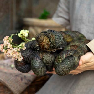 Può includere: Diverse matasse di filato tinto a mano in tonalità scure di verde bosco, bronzo e marrone intenso con una lucentezza sottile. Il filato è raggruppato e decorato con un piccolo mazzo di fiori rosa chiaro e color crema e un cartellino di carta beige.