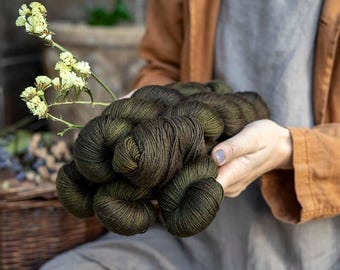 May include: A bundle of dark olive green yarn skeins with a subtle sheen, held alongside a delicate sprig of small, pale yellow dried flowers. The yarn features a deep, earthy tone with visible fiber texture and a tightly twisted structure.