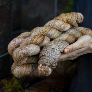 May include: A skein of hand-dyed yarn in shades of beige, brown, and tan. The yarn is held in a person's hands, showcasing its soft texture and color variations. Ideal for knitting or crochet projects.
