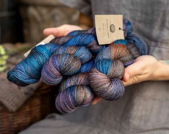 May include: Multiple skeins of hand-dyed yarn featuring a variegated color palette of deep blue, purple, and copper brown tones. A rectangular tan paper tag is attached to the yarn, displaying the text Hippie Fiber and a small heart icon.