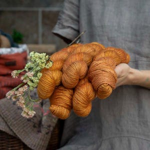 Puede incluir: Una colección de madejas de hilo de color naranja vibrante sujetas con un pequeño ramo de flores secas de color verde claro y crema. El hilo tiene una textura suave y retorcida con un tono de color cálido y saturado en un grupo.
