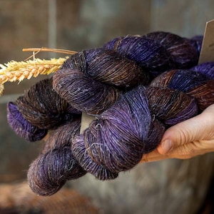 Puede incluir: Una madeja de hilo teñido a mano con una paleta de colores variados en morado intenso y marrón oscuro. El hilo tiene un brillo sutil y está retorcido en un paquete. Una pequeña etiqueta de papel rectangular de color marrón claro está unida al hilo y una espiga de trigo seca está insertada en las fibras.