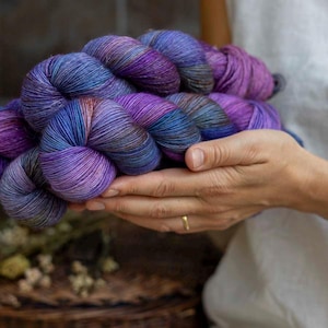 Può includere: Una matassa di filato tinto a mano caratterizzata da una tavolozza di colori variegati nei toni del viola intenso, del violetto vibrante e del blu scuro. Il filato è attorcigliato in un fascio a più anelli, con una consistenza morbida e fibrosa.