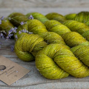 Puede incluir: Varias madejas de hilo teñido a mano en tonos vibrantes de verde lima y oliva con una apariencia texturizada y jaspeada. Se incluye una etiqueta de papel marrón con el texto "HIPPIE FIBRES" y un pequeño icono de corazón impreso.