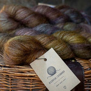 Puede incluir: Varias madejas de hilo teñido a mano en tonos variados de marrón, verde oliva y morado apagado. Se adjunta una etiqueta de papel rectangular de color marrón claro con el texto HIPPIE | FIBRES, una ilustración de ovillo y un corazón.