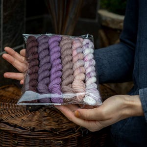May include: A clear plastic package containing a collection of yarn skeins in various shades of purple, brown, and pink. The yarn is arranged in a row, with each skein tightly wound. The package is held in a person's hands, with a wicker basket in the background.