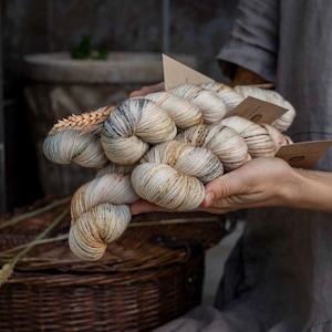 Puede incluir: Una colección de madejas de hilo teñidas a mano en un tono blanquecino neutro, con sutiles motas de color marrón, bronceado y gris carbón a lo largo de las fibras. Cada madeja está retorcida e incluye una pequeña etiqueta de papel marrón claro.