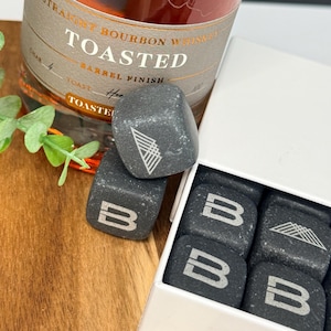 May include: A set of gray whiskey stones with white logos, displayed in a white box next to a bottle of "Toasted" bourbon whiskey. The stones have the letter "B" and a geometric design. The bottle has a brown label.