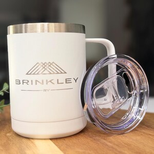May include: White travel mug with a silver rim and handle. The mug has the text "BRINKLEY RV" printed in silver. A clear plastic lid is next to the mug. The mug is on a wooden surface.