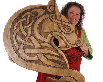 Op de afbeelding: Een houten carving van een wolfskop met ingewikkelde Keltische knoopwerk. De carving is gekleurd in tinten bruin en wordt vastgehouden door een persoon die een rode jurk en een groen shirt draagt.