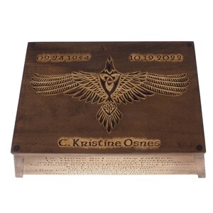 May include: A wooden box with a carved raven design and the inscription "C. Kristine Osnes". The dates "09.24.1954" and "10.19.2029" are also engraved on the box. The box has a quote on the bottom that reads "Lo, there I see my father...Where the brave may live forever."