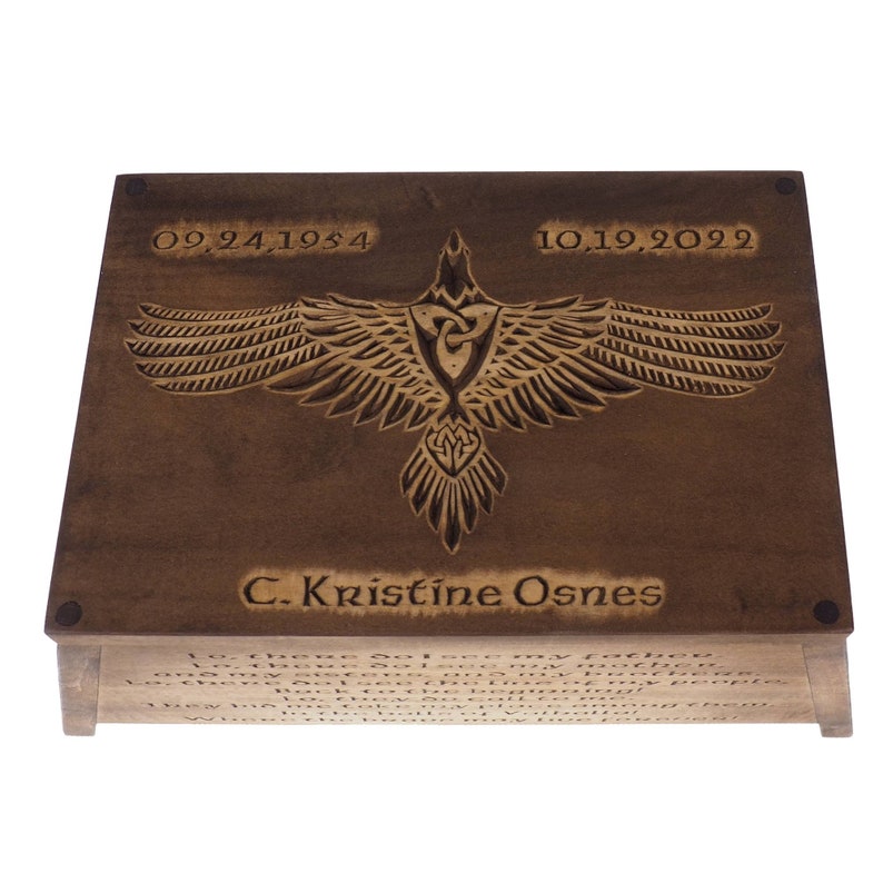 May include: A wooden box with a carved raven design and the inscription "C. Kristine Osnes". The dates "09.24.1954" and "10.19.2029" are also engraved on the box. The box has a quote on the bottom that reads "Lo, there I see my father...Where the brave may live forever."