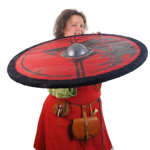 May include: A woman in a red dress holds a large, round shield with a black rim and a red center. The shield has a black design on it and a metal boss in the center.