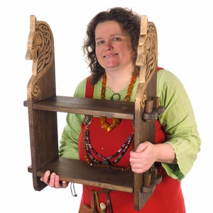 Könnte beinhalten: Ein Holzregal mit zwei Regalbrettern, die an jeder Seite mit einem Pferdekopf-Design verziert sind. Das Regal ist dunkelbraun und hat eine natürliche Holzoptik.