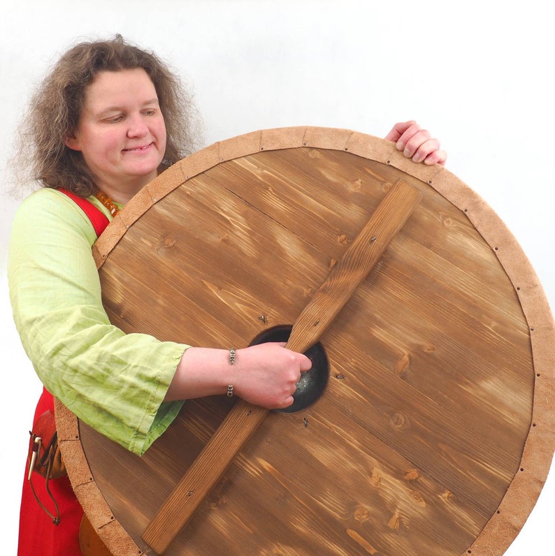 May include: A woman holds a large, round wooden shield with a leather trim. The shield has a wooden handle and a circular opening in the center.