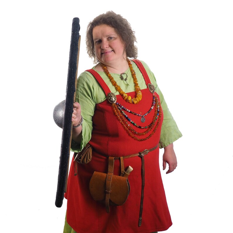 May include: A woman wearing a red dress with a green tunic over it, holding a round shield. She is wearing a necklace with amber beads and a silver pendant.
