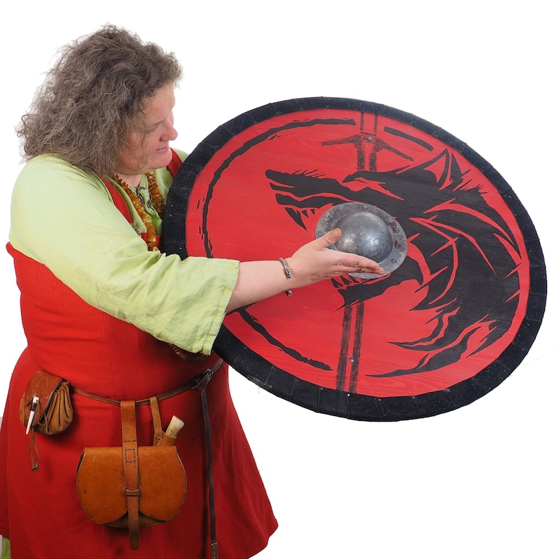May include: A woman in a red dress holds a round shield with a black wolf design on a red background. The shield has a metal boss in the center.