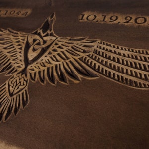 May include: A brown wooden plaque with a carved image of a stylized bird with outstretched wings. The bird has a Celtic knot design on its chest. The dates "09.24.1954" and "10.19.2022" are carved into the wood.