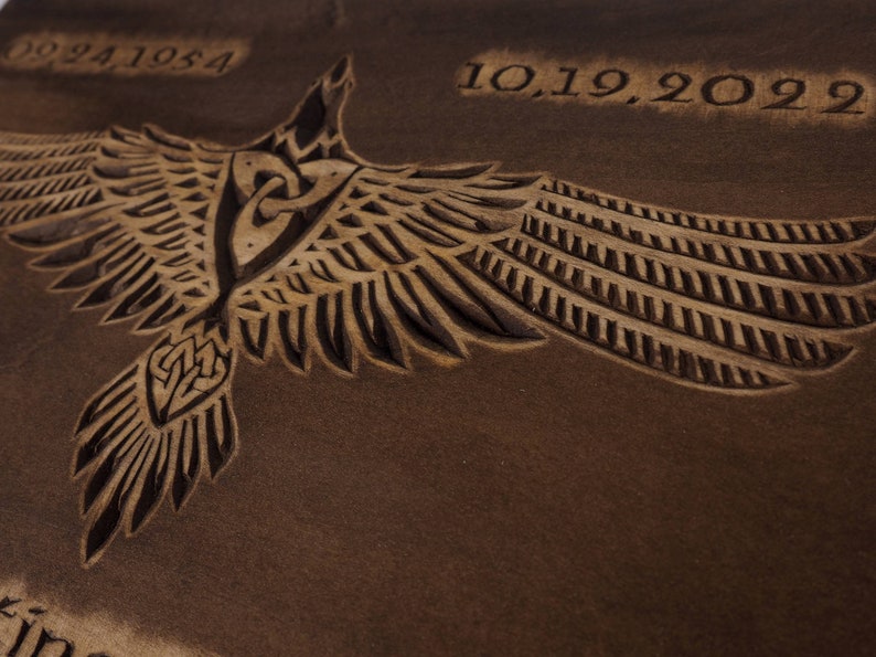 May include: A brown wooden plaque with a carved image of a stylized bird with outstretched wings. The bird has a Celtic knot design on its chest. The dates "09.24.1954" and "10.19.2022" are carved into the wood.