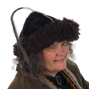 Può includere: Un cappello nero tradizionale con finiture in pelliccia e un lungo ornamento a forma di capelli. Il cappello presenta finiture in pelliccia marrone scuro e una parte superiore nera, con cuciture bianche lungo i bordi. Parte di un costume storico o culturale.