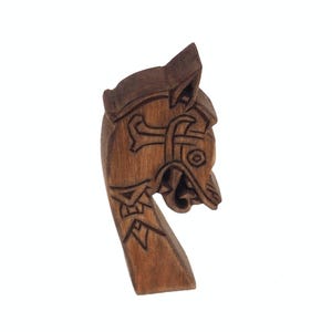 Könnte beinhalten: Eine geschnitzte Wikinger-Schiffs-Bugfigur in Brauntönen. Die Bugfigur zeigt einen stilisierten Tierkopf mit aufwendigen Schnitzereien, darunter ein kreuzartiges Design und ein offener Mund. Das Holz hat eine natürliche Maserung.