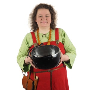 Puede incluir: Una mujer con un vestido rojo y verde con un collar de ámbar sostiene una olla de metal negro con asa.