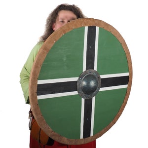 Puede incluir: Escudo redondo de madera con fondo verde y diseño de cruz negra y blanca. El escudo tiene un borde de cuero marrón y un umbo metálico en el centro. Réplica histórica.
