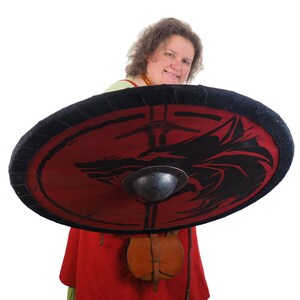 May include: A woman in a red and green tunic holds a round shield with a black wolf design on a red background. The shield has a black rim and a metal center boss.
