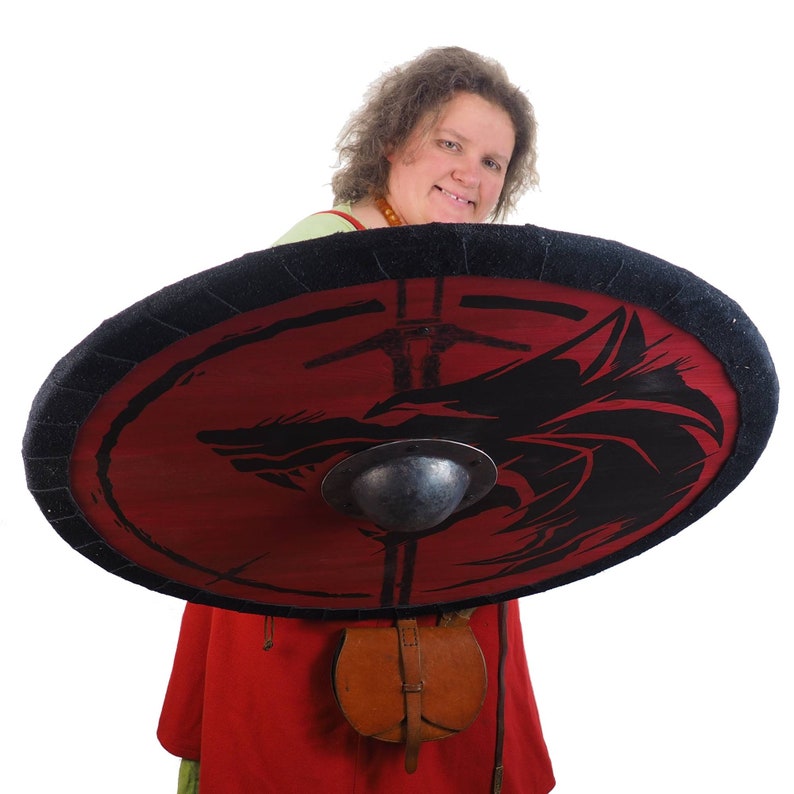 May include: A woman in a red and green tunic holds a round shield with a black wolf design on a red background. The shield has a black rim and a metal center boss.