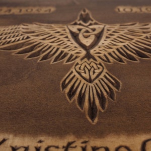 May include: A brown wooden plaque with a carved design of a bird with outstretched wings. The bird has a Celtic knot design in its chest and tail. The plaque also has the dates "1992 1984 1992" carved into it.