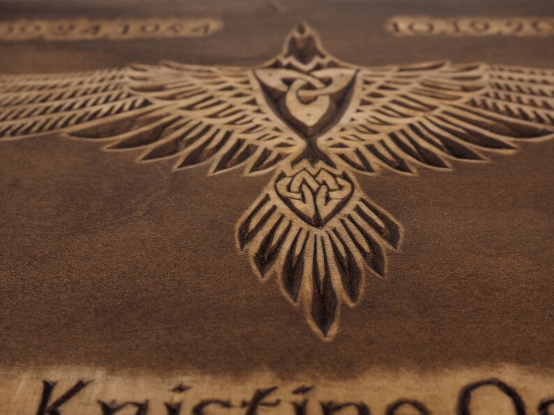 May include: A brown wooden plaque with a carved design of a bird with outstretched wings. The bird has a Celtic knot design in its chest and tail. The plaque also has the dates "1992 1984 1992" carved into it.