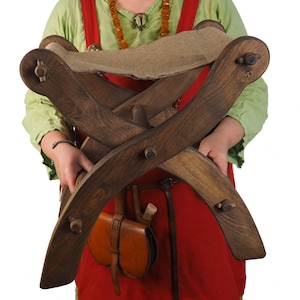 May include: A dark brown wooden folding stool with a tan fabric seat. The stool is held by a person wearing a green shirt and red apron. The stool is approximately 18 inches long.
