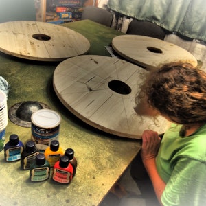 May include: Four wooden round table tops with a hole in the center, sitting on a green surface.  A person is working on one of the table tops.  There are several small bottles of paint and a can of stain on the surface.
