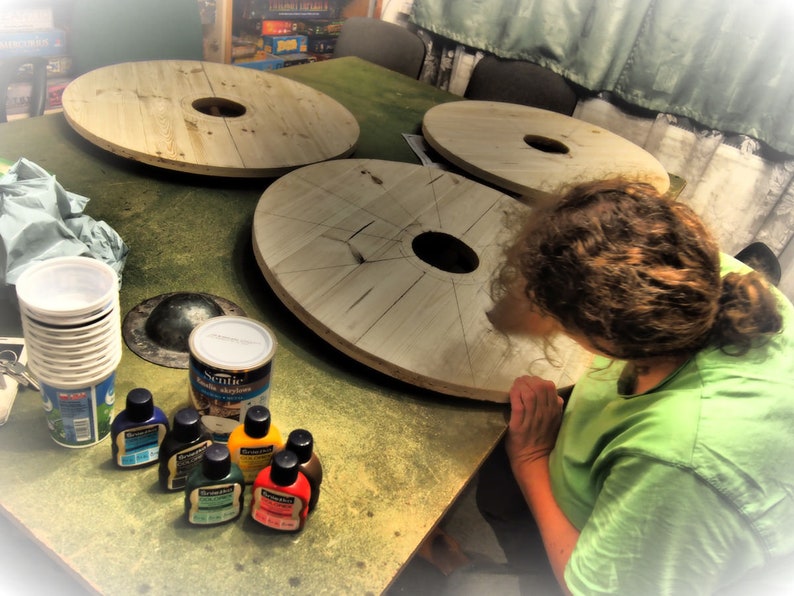 May include: Four wooden round table tops with a hole in the center, sitting on a green surface.  A person is working on one of the table tops.  There are several small bottles of paint and a can of stain on the surface.