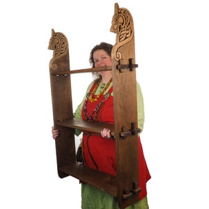 Könnte beinhalten: Ein Holzregal mit zwei Ablagen und dekorativen Schnitzereien von Tierköpfen. Die Person hält das Regal. Das Holz hat einen warmen, braunen Farbton. Das Regal ist im Wikinger-Stil gestaltet. Maße in Zentimetern.