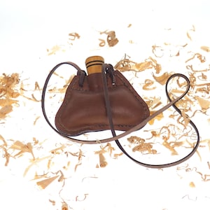 May include: A brown leather pouch with a drawstring closure and a wooden stopper. The pouch is hanging from a brown leather strap. The pouch is surrounded by wood shavings.