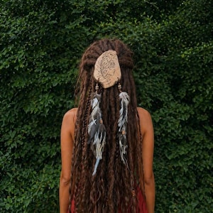 Puede incluir: Accesorio para el cabello de madera con un rostro grabado y detalles de plumas. La pieza para el cabello está sujeta a largas rastas marrones. Dos mechones de plumas cuelgan, agregando un toque bohemio. El fondo es un exuberante arbusto verde.